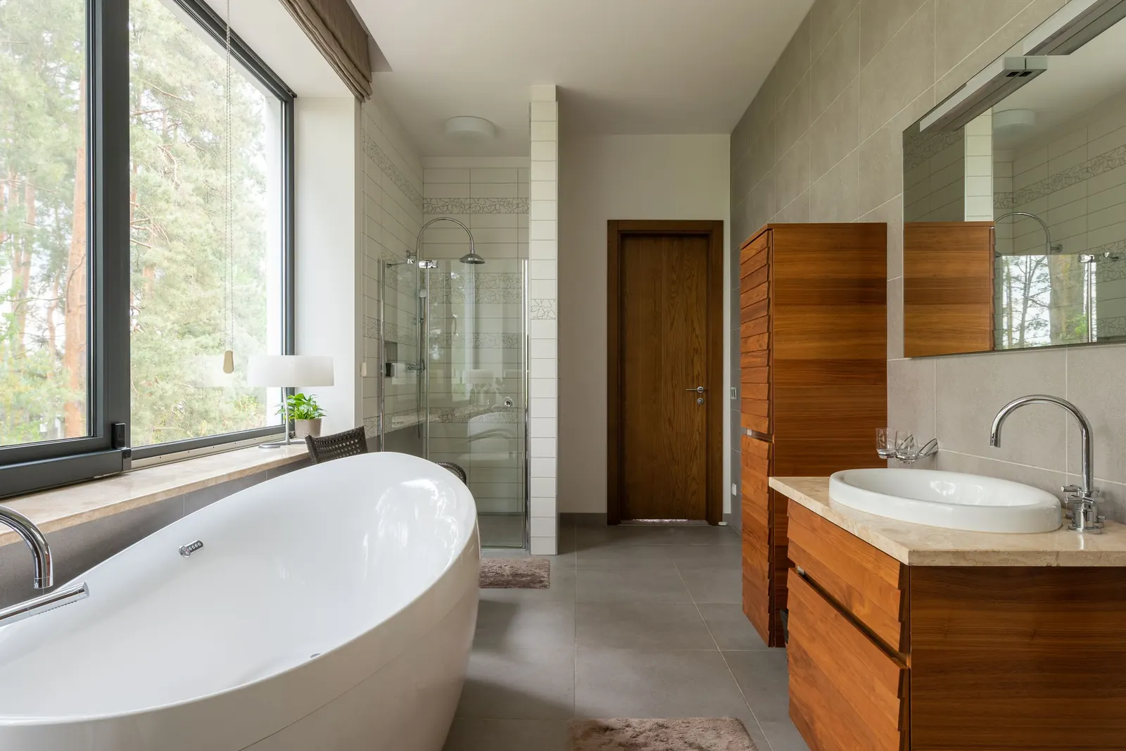 Bright bathroom with a freestanding bath in front of a large window facing the woods, a glass walled shower and a basin on a wooden unit